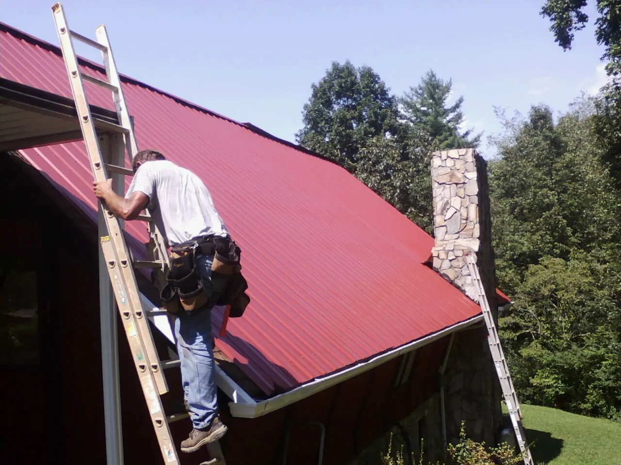 Roofer on a ladder beside a red metal roof and stone chimney — Soffit & Fascia Repair by Roofing Brothers in Sunol